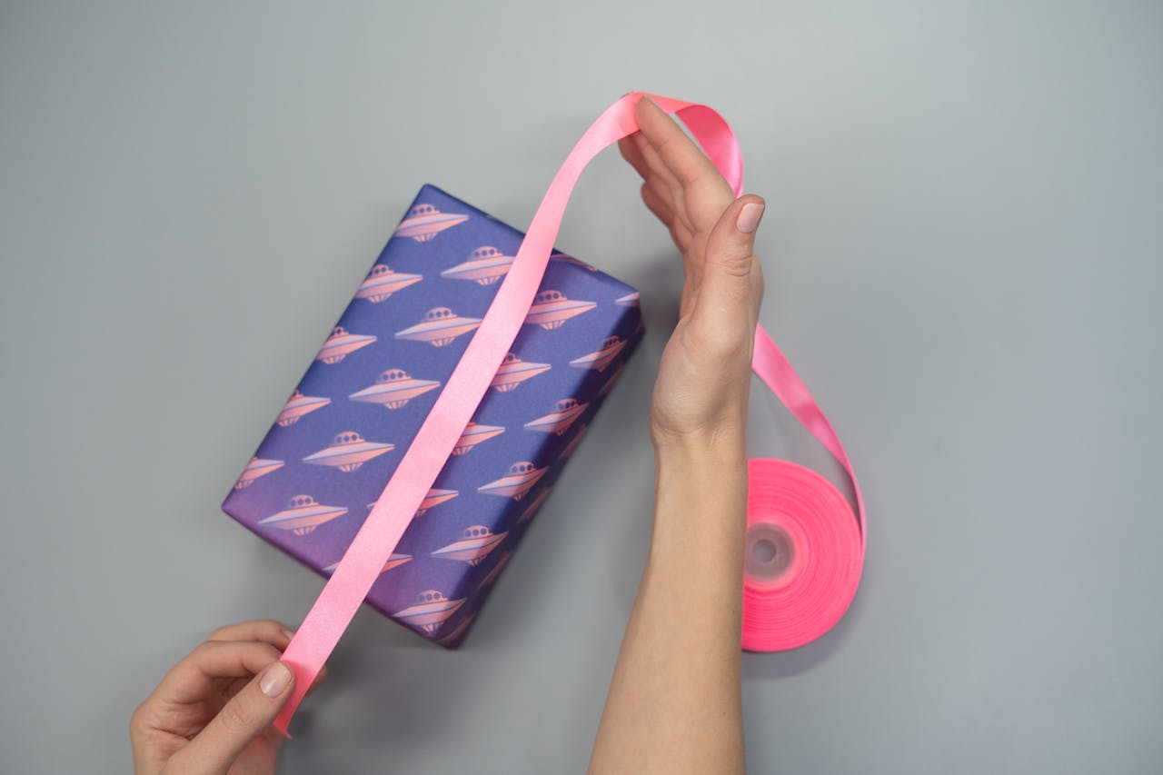 A close-up of hands wrapping a gift box with pink ribbon and UFO-themed paper.