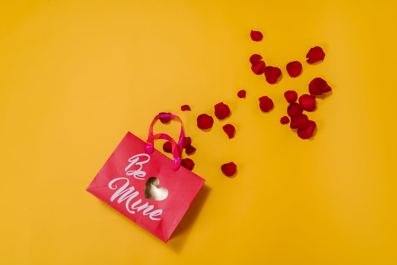 Pink gift bag with 'Be Mine' text on yellow background, surrounded by red rose petals, perfect for Valentine's Day.